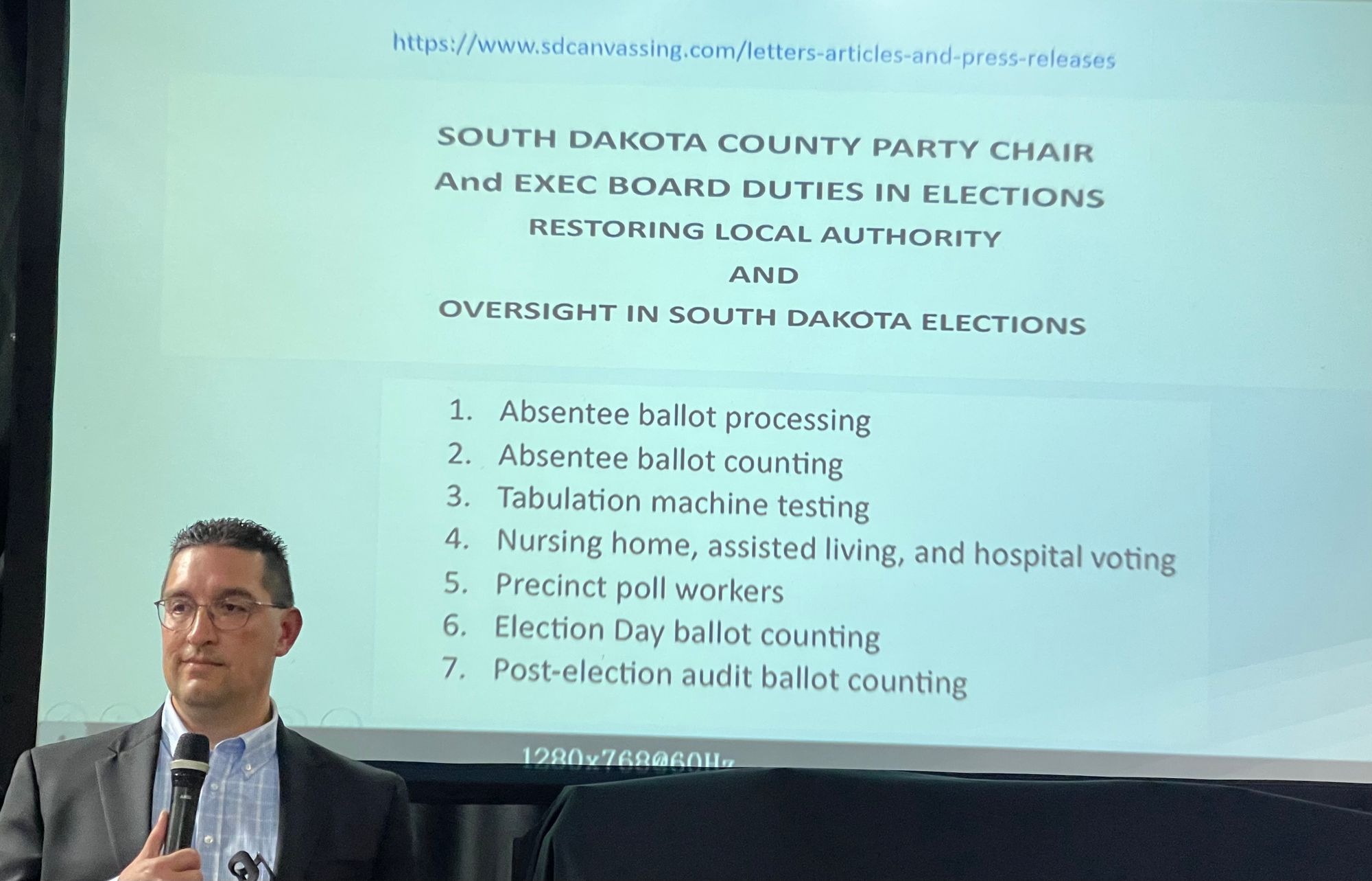 Rick Weible holds a microphone while standing in front of a screen that is showing a presentation on elections.