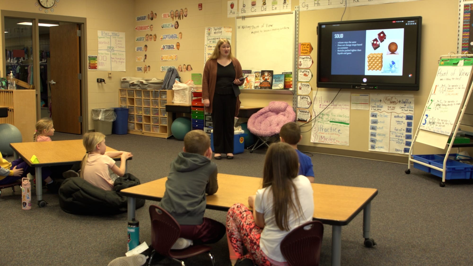 A teacher leads a lesson at an elementary school classroom with kids looking on.