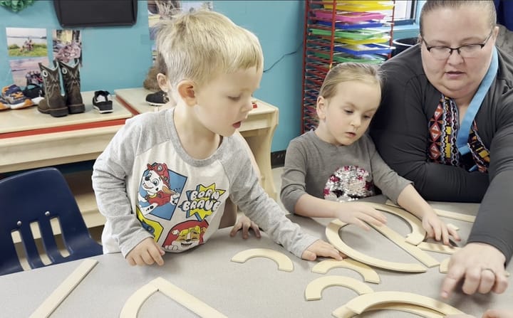 Preschool students in a classroom use wooden blocks to create letters as a teacher watches