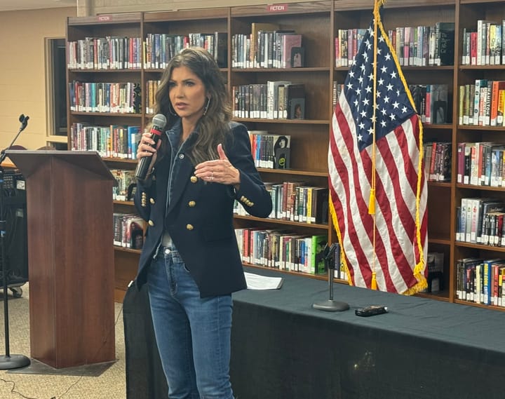 South Dakota Gov. Kristi Noem speaks in front of a wall of books with the American flag in the background