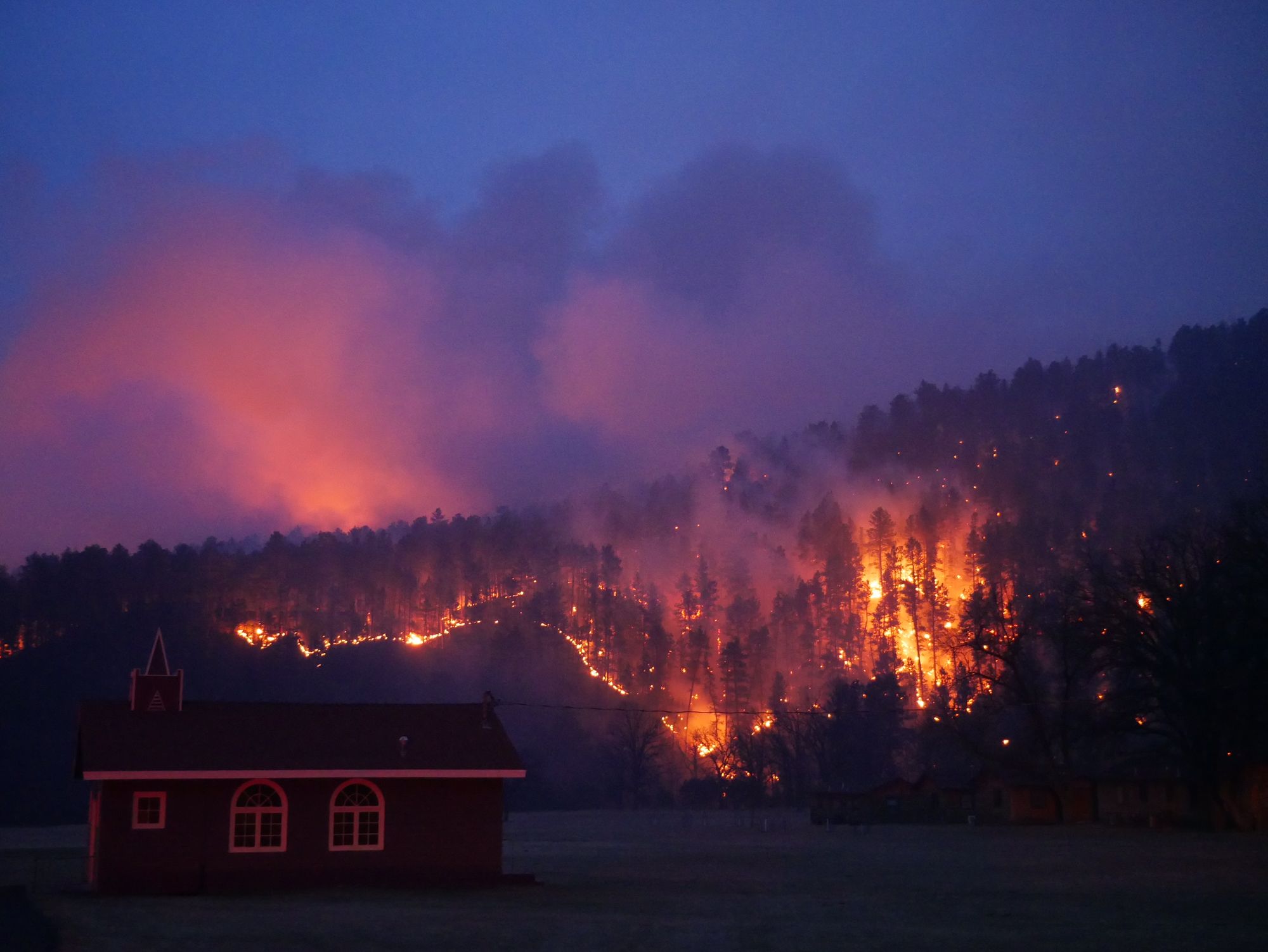 A cabin at night while a forest fire burns in the hills in the background